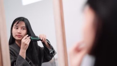 Asian woman using a hair straightener to achieve smooth hair in front of a large mirror after washing and styling at home. Showcase the beauty routine for enhanced sales. High quality 4k footage
