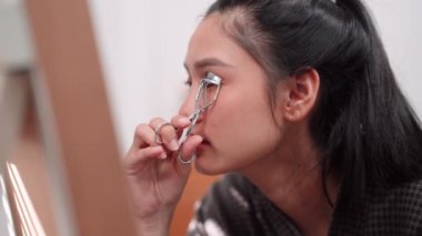 Asian woman using an eyelash curler in front of a large mirror after bathing and styling her hair at home. Showcase her beauty routine. High quality 4k footage