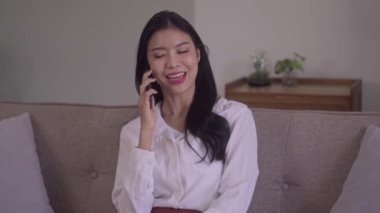 Happy young Asian woman talking on mobile smartphone sit sofa indoors at modern home. High quality 4k footage