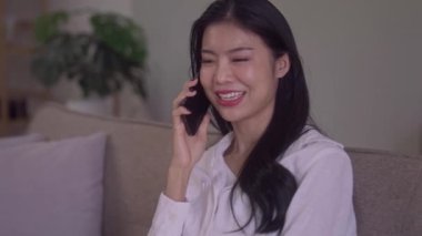 Happy young Asian woman talking on mobile smartphone sit sofa indoors at modern home. High quality 4k footage
