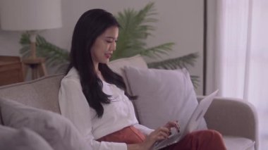 Young business freelance Asian Woman watching laptop to checking social media, surfing, doing online ecommerce shopping while lying on the sofa when relax in living room at home
