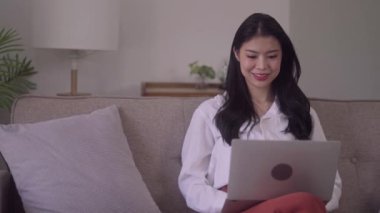 Young business freelance Asian Woman watching laptop to checking social media, surfing, doing online ecommerce shopping while lying on the sofa when relax in living room at home