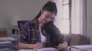 Young Asian female graphic designer at desk sketching or drawing design on white notebook at workshop. High quality 4k footage