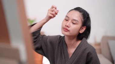 An Asian woman applying face cream to nourish her skin in front of a large mirror after washing and styling at home. Highlight the skincare routine. High quality 4k footage