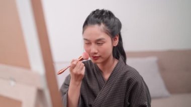 Asian woman applying bronzer on her face in front of a large mirror after bathing and styling her hair at home. Capture her beauty routine for increased sales and skincare promotion