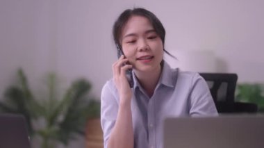 Genç Asyalı iş kadını cep telefonuyla konuşuyor ya da ev ofisinde çalışırken kurumsal iş sorununu çözmek için akıllı telefon kullanıyor. Yüksek kalite 4k görüntü
