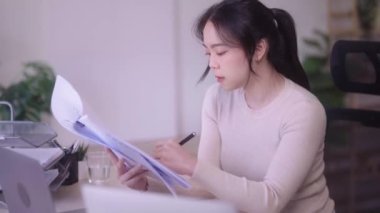 Young Asian woman hard working with documents at her using laptop computer with analytical charts and graphs at her home, work from home concept. High quality 4k footage