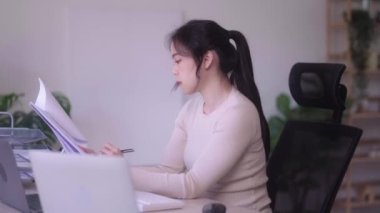 Young Asian woman hard working with documents at her using laptop computer with analytical charts and graphs at her home, work from home concept. High quality 4k footage