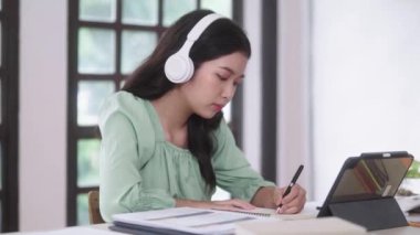 Genç Asyalı üniversite öğrencisi kadınlar evdeki oturma odasında dijital tabletle ve kulaklıkla e-öğrenim görüyorlar, uzaktan eğitim kavramı. Yüksek kalite 4k görüntü