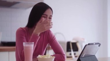 Neşeli, genç Asyalı, günlük kıyafetli genç bayan mutfak tezgahında dinlenirken portatif tablette film izlerken abur cubur yiyor. Yüksek kalite 4k görüntü