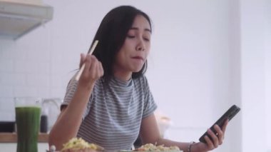 Asya eriştesi yiyen neşeli güzel bir Asyalı kadın öğle yemeği arasında yemek çubuğu tutuyor ve akıllı telefonuyla yazı yazıyor. 