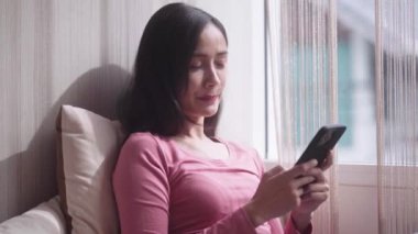 Neşeli, güzel, güzel, Asyalı bir kadın evdeki pencere eşiğinde oturuyor ve telefonuyla mesajlaşıyor ve evdeki pencerenin yanındaki internet uygulamasını kullanıyor. Yüksek kalite 4k görüntü