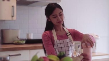 Genç Asyalı bir kadın sağlıklı bir içecek ile karıştırıcıyı karıştırıyor sağlıklı bir içecek için evdeki mutfağındaki tezgahta kahvaltıda bir bardakta. Yüksek kalite 4k görüntü