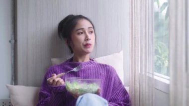Asyalı sağlıklı kadın salata kasesi tutuyor ve öğle yemeğinde kilo verme diyeti ve pencerenin yanında sağlıklı beslenme ve sağlıklı gıda konsepti ile temiz sebzeler yiyor. Yüksek kalite 4k görüntü