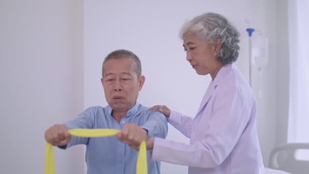 アジアの女性の物理療法士の介護者は伸縮性がある抵抗バンドが付いている物理的な部屋の成熟したアジアの男性灰色の毛の患者と助け 運動します — ストック動画