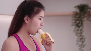 Spor giyimli genç bir Asyalı kadın mutfak içinde muz yiyor, meyve diyeti ve sağlıklı spor kilo verme kavramının tadını çıkarıyor. Yüksek kalite 4k görüntü