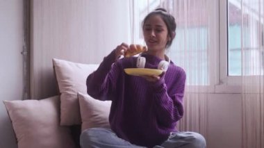 Genç, çekici, gülümseyen Asyalı kadın sabahın erken saatlerinde kruvasan yiyor, pencerenin kenarında oturuyor. Yüksek kalite 4k görüntü