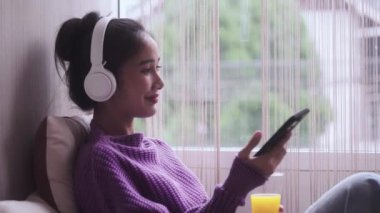Pencerelerin yanında oturan, portakal suyu içen, cep telefonu ve kulaklıkla müzik dinleyen mutlu genç Asyalı kadın. Yüksek kalite 4k görüntü