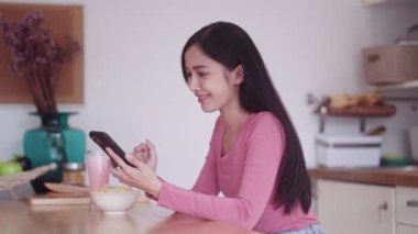 Neşeli, genç Asyalı, günlük kıyafetli, akıllı telefondan film izlerken evde mutfak tezgahında rahatlarken abur cubur yiyen genç bir kız. Yüksek kalite 4k görüntü