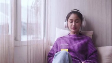 Pencerelerin yanında oturan, portakal suyu içen, cep telefonu ve kulaklıkla müzik dinleyen mutlu genç Asyalı kadın. Yüksek kalite 4k görüntü