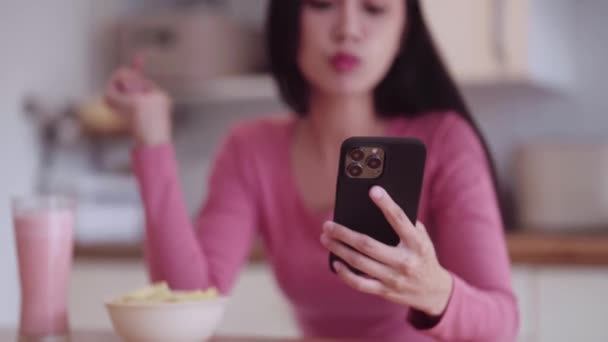 カジュアルな衣装で陽気な若いアジアの女性ティーンエイジャーは 自宅のキッチンのカウンターでリラックスしながら スマートフォンでフィルムを見ているときにジャンクファストフードスナックチップを食べます 高品質の4K映像 — ストック動画
