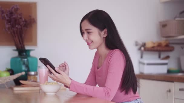 カジュアルな衣装で陽気な若いアジアの女性ティーンエイジャーは 自宅のキッチンのカウンターでリラックスしながら スマートフォンでフィルムを見ているときにジャンクファストフードスナックチップを食べます 高品質の4K映像 — ストック動画