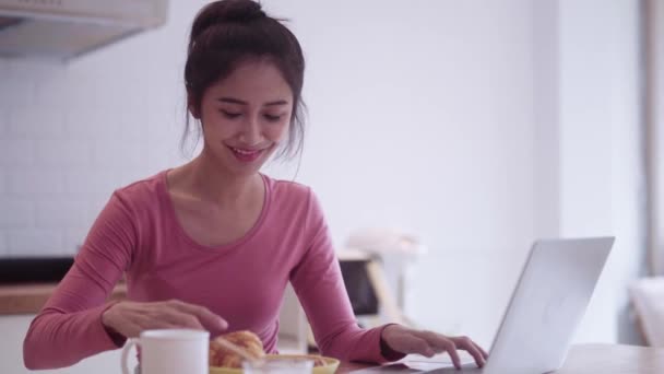 若い幸せなアジアフリーランス女性は朝食のためにクロワッサンを食べて キッチンカウンターでラップトップで作業しながらコーヒーを飲んで 自宅で働くことを楽しんでいます — ストック動画