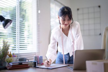 Boynuna mezuralar takan Asyalı genç tüccar kadınlar ve internet üzerinden siparişleri kontrol ediyorlar. İç işlerindeki online alışveriş işlerinde çalışırken not alıyorlar..