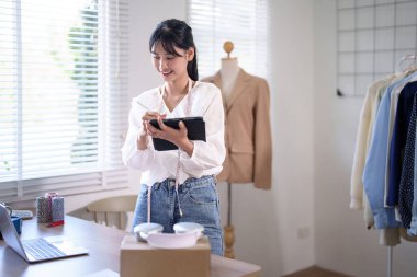 Boynuna mezura takan Asyalı genç tüccar kadınlar dijital tabletle müşteri bilgilerini yazarken dizüstü bilgisayardan online siparişleri kontrol ediyor ve çevrimiçi alışveriş işlerinde çalışıyor..