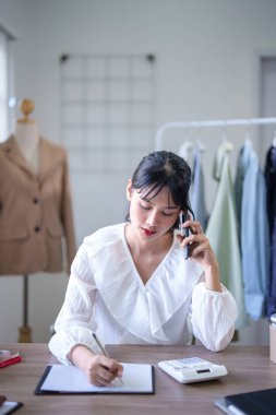 Genç Asyalı tüccar kadınlar akıllı telefonu müşteri bilgileri ve kıyafet siparişleri hakkında konuşmak için kullanıyorlar. İç işlerindeki çevrimiçi alışveriş işlerinde çalışırken ve nakliyat yaparken not alıyorlar..