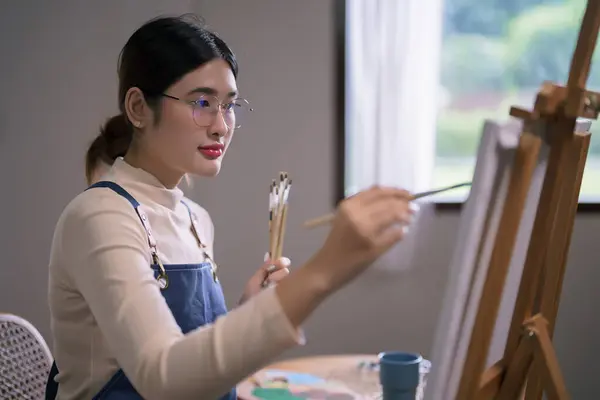 Young asian female artist hold paintbrush and use brush to drawing and painting artwork on canvas.