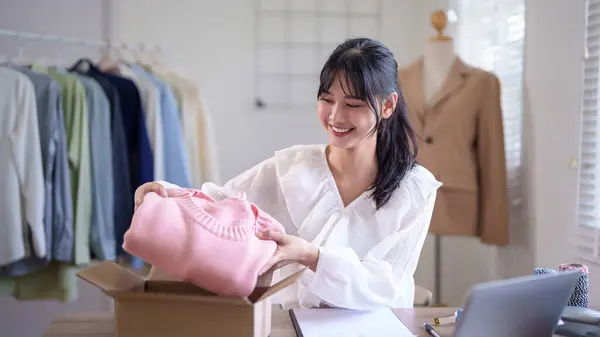 Young asian merchant women checking purchase online order on laptop and packaging clothes product in parcel to shipping delivery for client while working about online shopping business in home office.