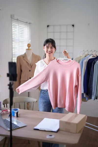 Young asian merchant women using smartphone to recording live streaming and selling clothes online in social media while working and shipping delivery for online shopping business in home office.