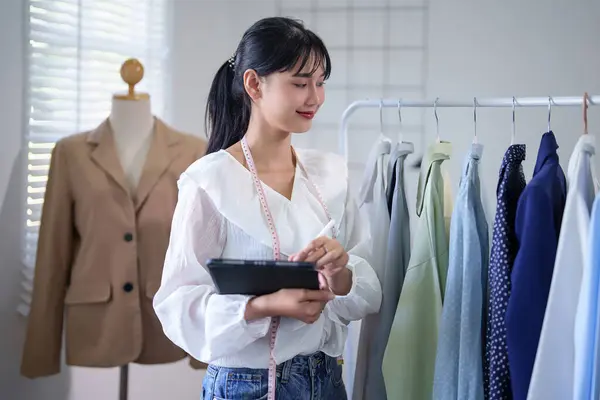 Young asian merchant women checking stock clothes product in store and customer online order information on digital tablet while taking notes and working about online shopping business in home office.