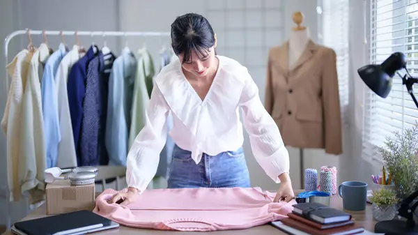 Young asian merchant women using measure tape to checking chest size details of fashion clothes on the table while taking notes in tablet and working about online shopping business in home office.