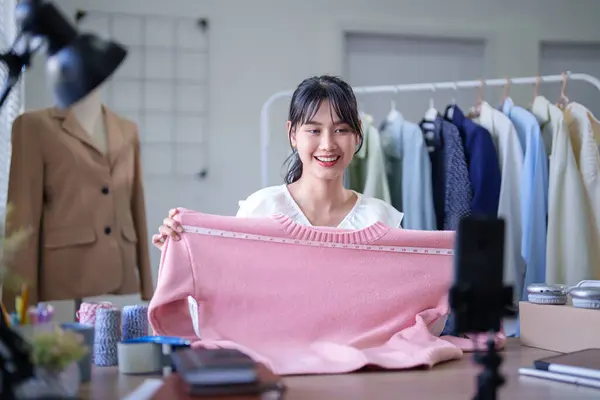Young asian merchant women using measure tape to checking size of sweater and selling clothes product while recording video for social media on smartphone and working about online shopping business.