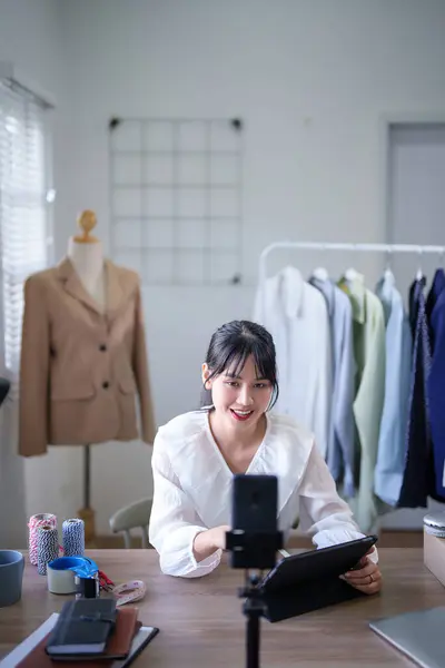 Young asian merchant women use smartphone to recording video and selling clothes product in social media while checking purchase orders in digital tablet and working about online shopping business.