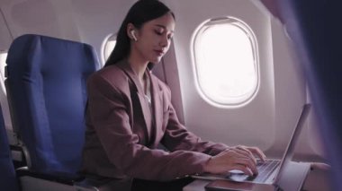 Asyalı bir iş kadını dizüstü bilgisayarı üzerinde çalışıyor, uçuş sırasında görevleri aktif olarak kaydediyor ve analiz ediyor. Ulaşım sırasında verimliliği maksimuma çıkarmaya kendini adadı.