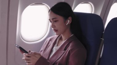 Asyalı iş kadını cep telefonuyla şarkı seçiyor ve uçakta seyahat ederken en sevdiği müziği kulaklıkla dinliyor.. 