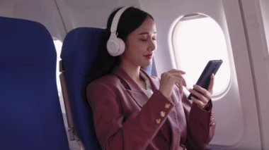 Asyalı iş kadını cep telefonuyla şarkı seçiyor ve uçakta seyahat ederken en sevdiği müziği kulaklıkla dinliyor.. 