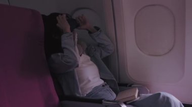 Asyalı kadın turist uykusundan uyanır, maskesini açar ve tatil günlerinde tatil yerine giderken gökyüzü manzarasının tadını çıkarmak için uçağın penceresinden dışarı bakar.