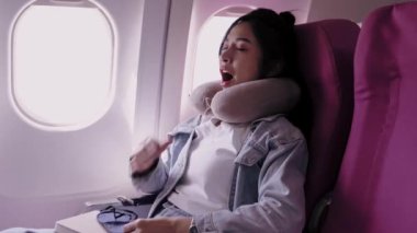 Asyalı kadın turist uykusundan uyanır, maskesini açar ve tatil günlerinde tatil yerine giderken gökyüzü manzarasının tadını çıkarmak için uçağın penceresinden dışarı bakar.