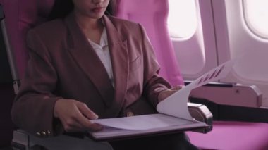 Asyalı bir iş kadını uçakta seyahat ederken iş belgelerini titizlikle analiz ediyor. O, uçuş zamanını en iyi şekilde değerlendirmek için tamamen işine daldı.
