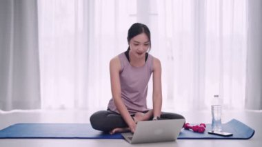 Antrenman kıyafeti giymiş Asyalı bir kadın evindeki yoga minderinin egzersiz bilgilerini bulmak için bilgisayarını kullanıyor. Sağlıklı bir yaşam tarzı ve evde yoga pratiği sergilemek için ideal.