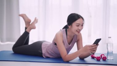 Antrenman kıyafeti giymiş Asyalı bir kadın, evinde yoga minderinde oturan bir arkadaşıyla akıllı telefonuyla sohbet ediyor. Aktif yaşam tarzlarını ve rahat bir yoga alanını sergilemek için mükemmel.