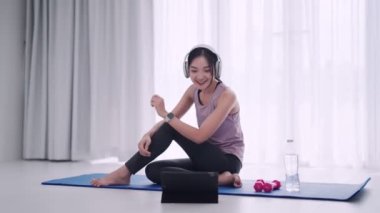 Antrenman kıyafeti giymiş Asyalı bir kadın evindeki yoga minderinde kulaklıkla müzik dinlemek için tablet kullanıyor. Formunu yükseltmek için mükemmel. 