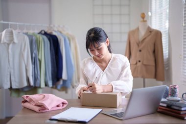 Genç Asyalı tüccar kadınlar çevrimiçi alışveriş işlerinde çalışırken karton kutuya adres yazmak için online siparişleri ve müşteri bilgilerini kontrol ediyorlar..