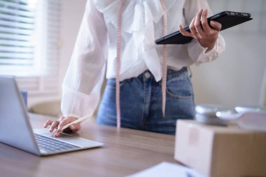 Genç Asyalı tüccar kadınlar online siparişleri ve müşteri bilgilerini kontrol etmek için dizüstü bilgisayarda veri yazarken tablete not alıyor ve merkez ofisteki çevrimiçi alışveriş işleri hakkında çalışıyor..