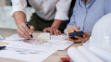 İki Asyalı mimar, model ve inşaat planı üzerinde çalışırken mimari plan projesinin çizimi hakkında tartışmak için hesap makinesi ve defter üzerine veri yazarlar..