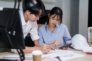 İki Asyalı mimar, model ve inşaat planı üzerinde çalışırken mimari plan projesinin çizimi hakkında tartışmak için hesap makinesi ve defter üzerine veri yazarlar..